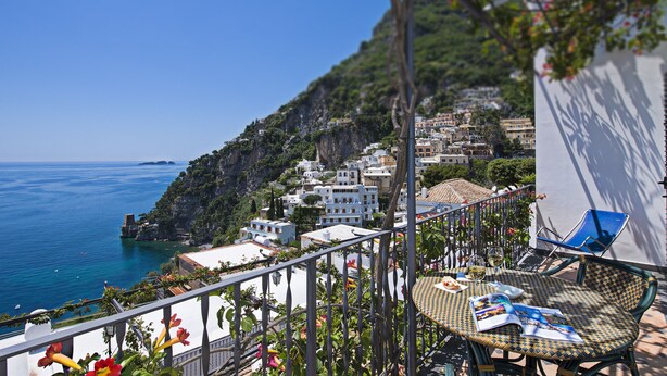 A balcony with a view of a coastal town, a table with a magazine, and a chair.