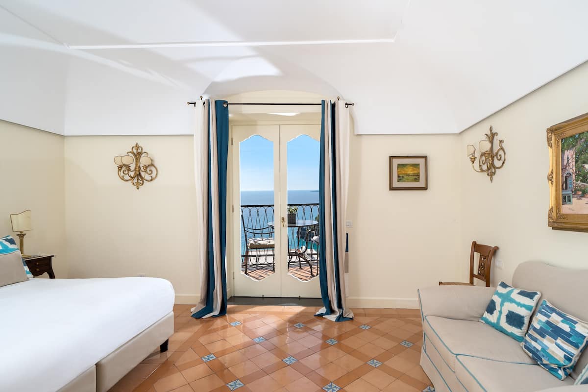 A bedroom with a bed, a sofa, a view through a door, and a tiled floor.