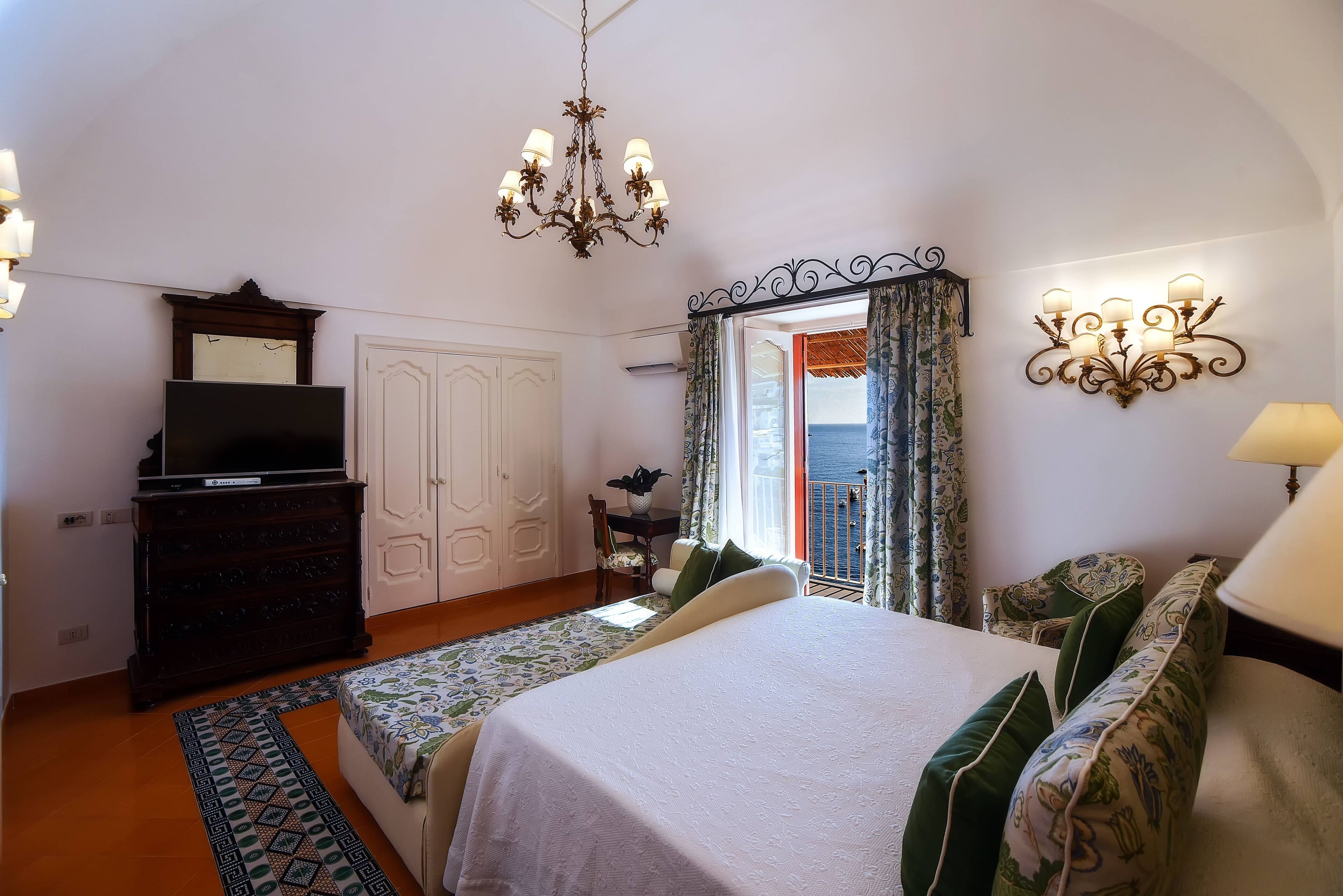 A bedroom with a bed, dresser, TV, and a view of the sea through a window.