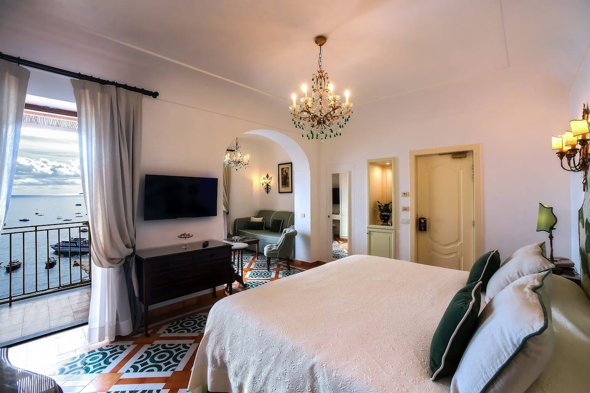 Una habitación de hotel con una cama grande, televisión, balcón con vistas a barcos y un candelabro.
