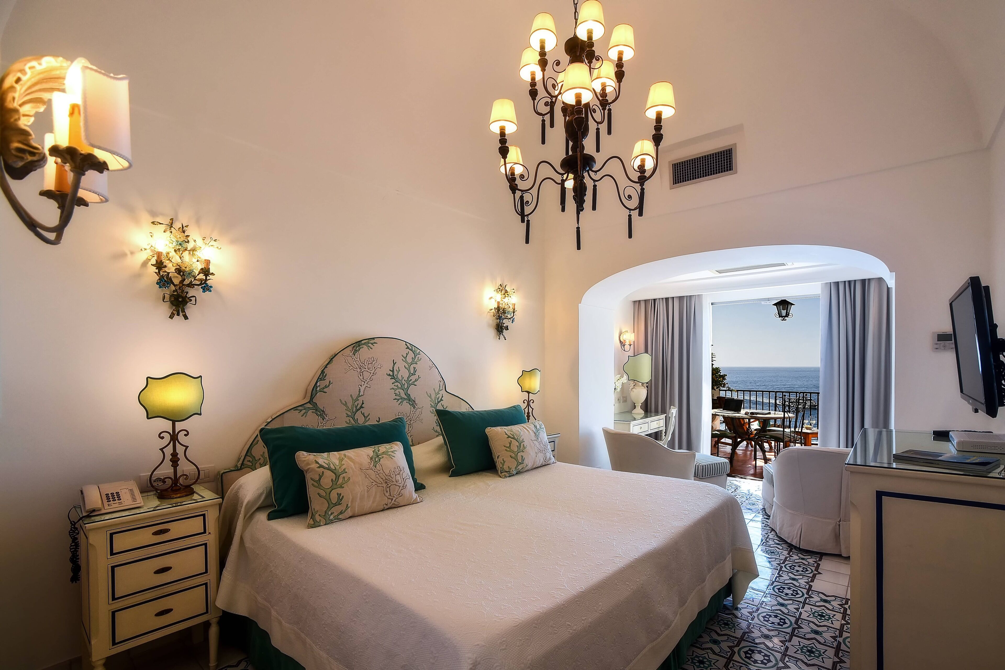 A hotel room with a bed, bedside lamps, a desk, and a view of the sea.