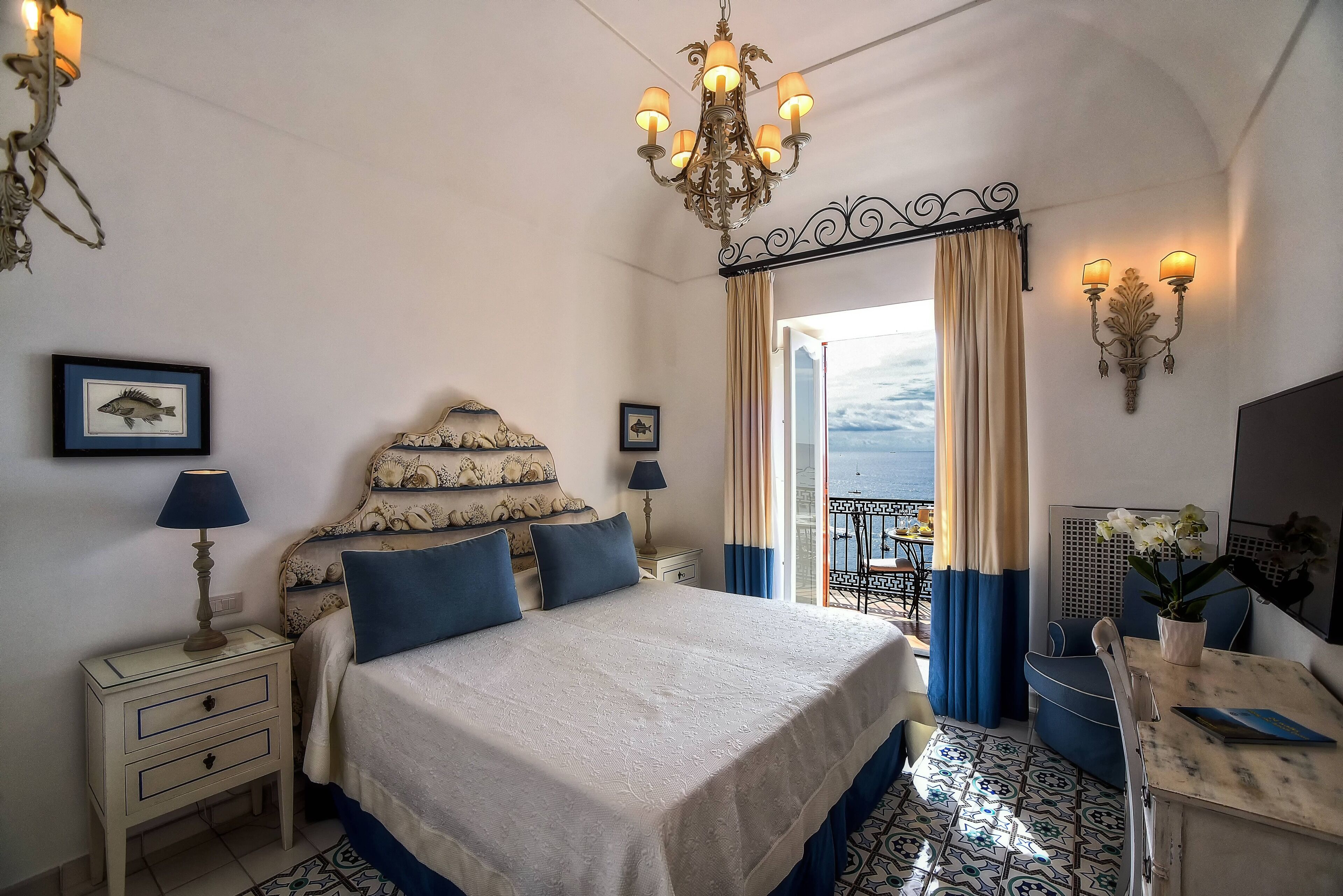 Ein Hotelzimmer mit Bett, Nachttischen, einer Kristallleuchtre, einem Balkon mit Vorhängen und Meerblick.