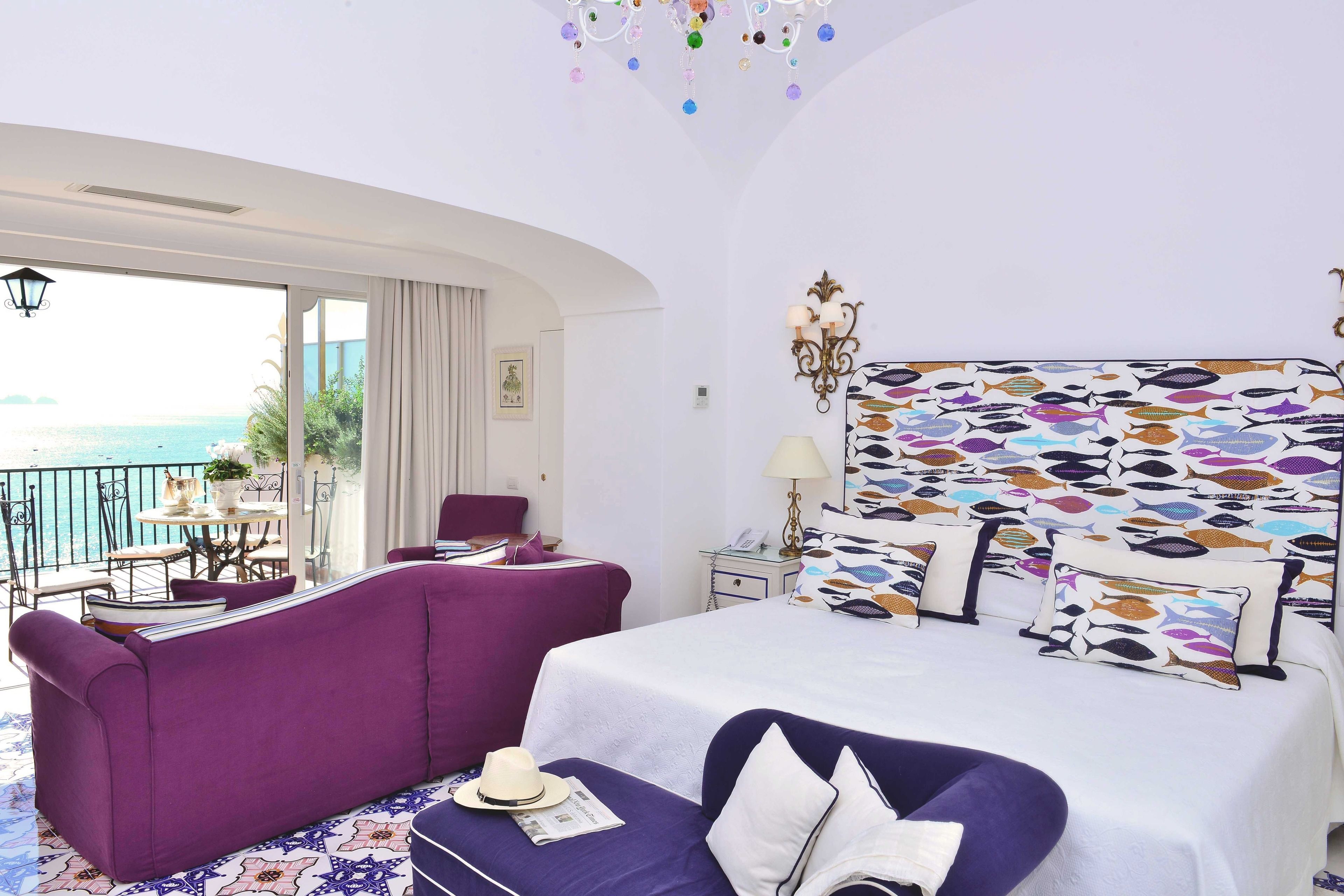 Habitación de hotel con una cama grande, un sofá morado, un balcón con vistas al mar y un cabecero decorado con motivos de peces.