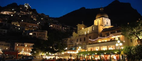 A coastal town at night with illuminated buildings, a prominent church with a dome, and a bustling waterfront area.