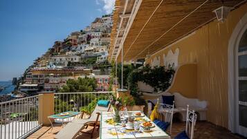 Un balcón con mesa dispuesta para comer, con vistas a un pueblo costero con edificios escalonados.