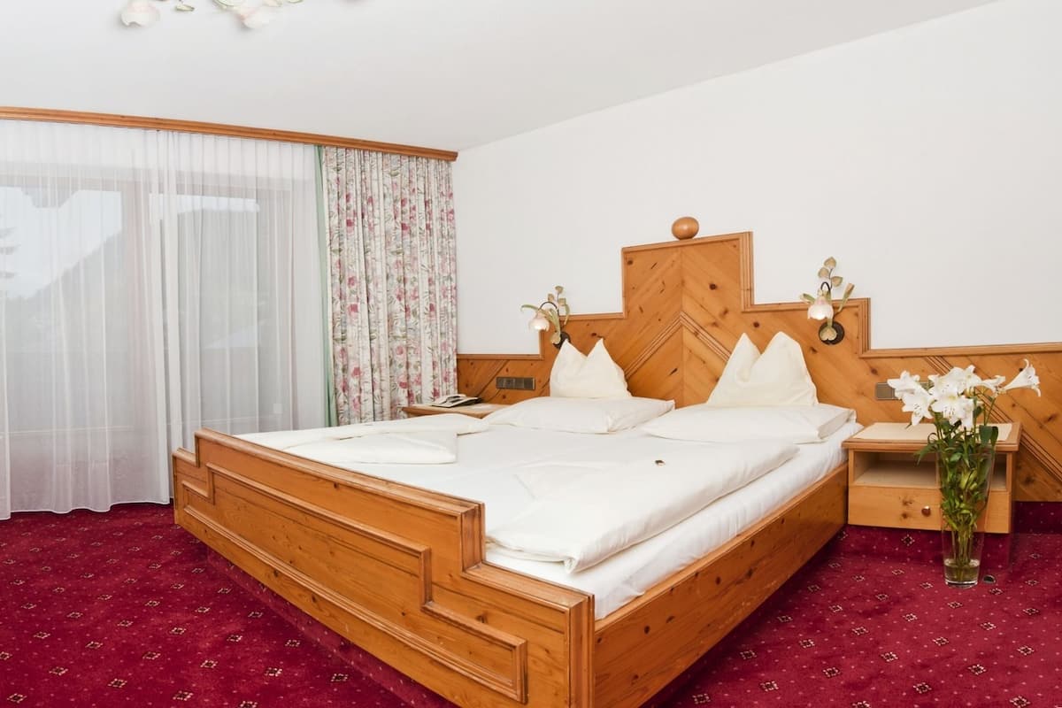 A hotel room with a wooden bed, a desk with a television, a small chair, and a large window with floral curtains.