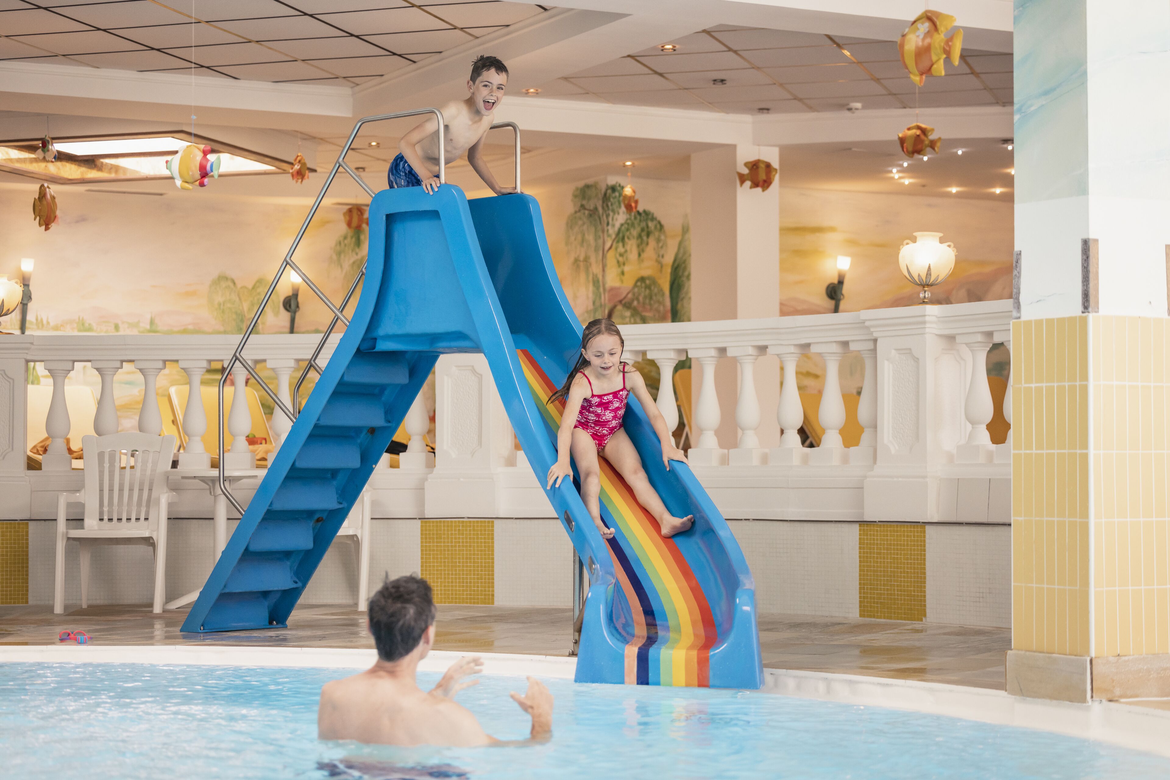 Un niño y un adulto en la cima de un tobogán azul con diseño de arcoíris, que desemboca en una zona de piscina con barandilla blanca y un mural en la pared.