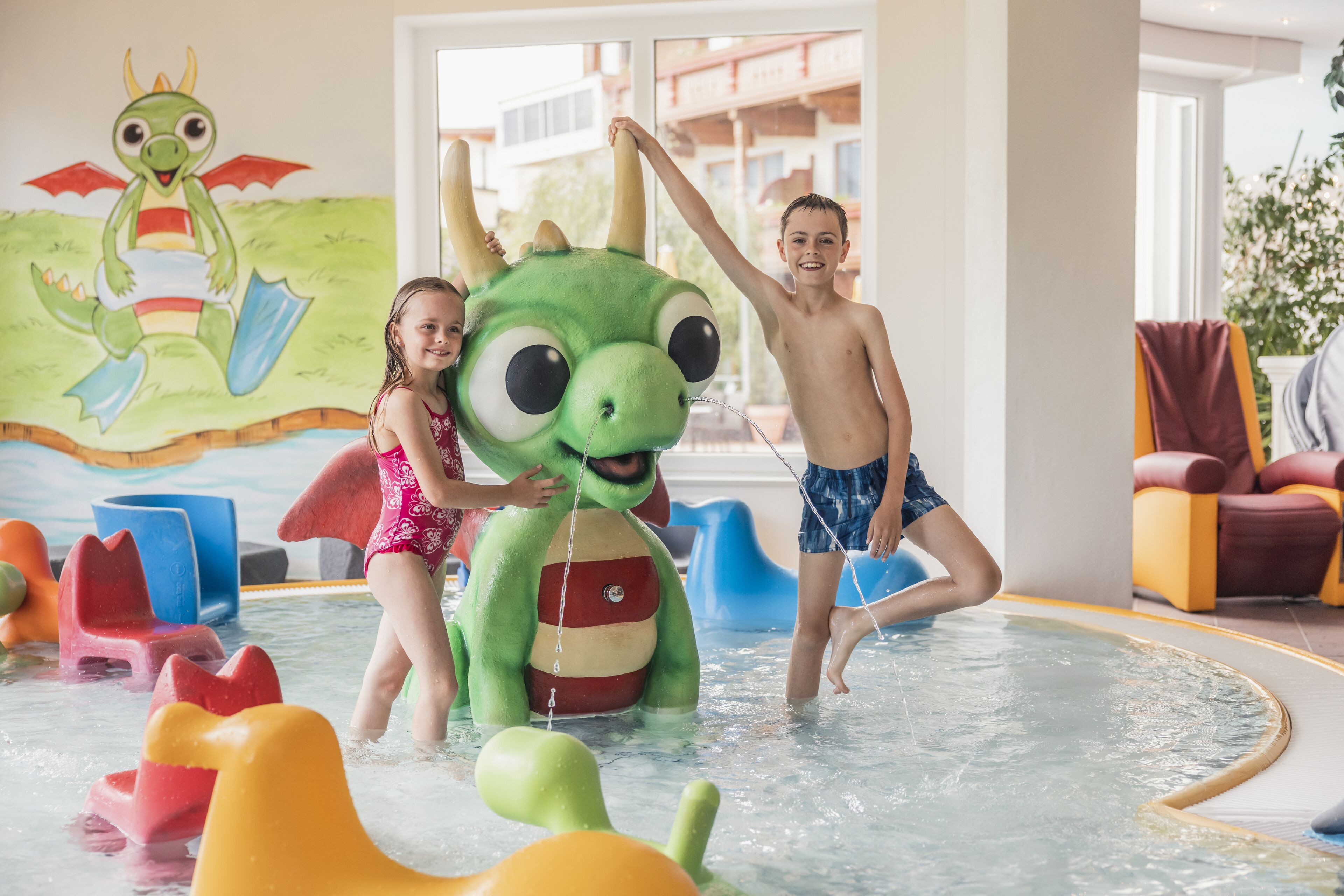 Area giochi con acqua, con un grande dinosauro gonfiabile verde e bambini che giocano.