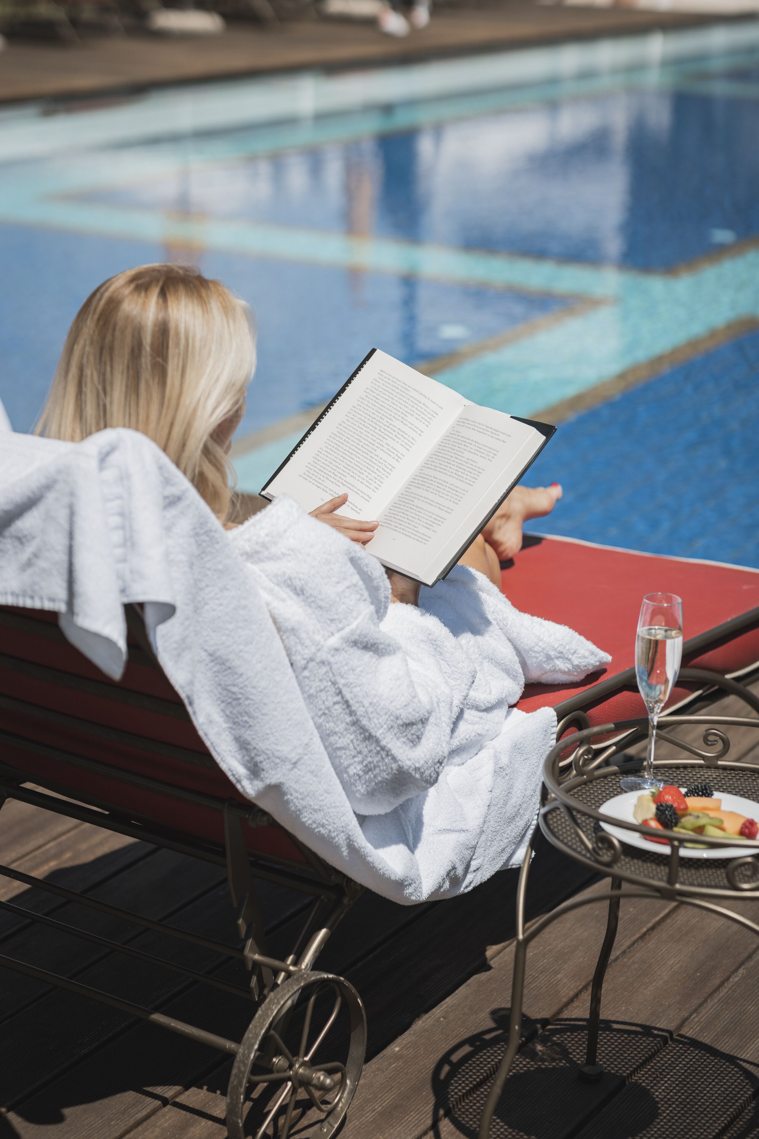 Une personne, vêtue d’un peignoir blanc, est assise près d’une chaise au bord de la piscine, un livre à la main, et a devant elle un verre de champagne et une assiette de fruits.