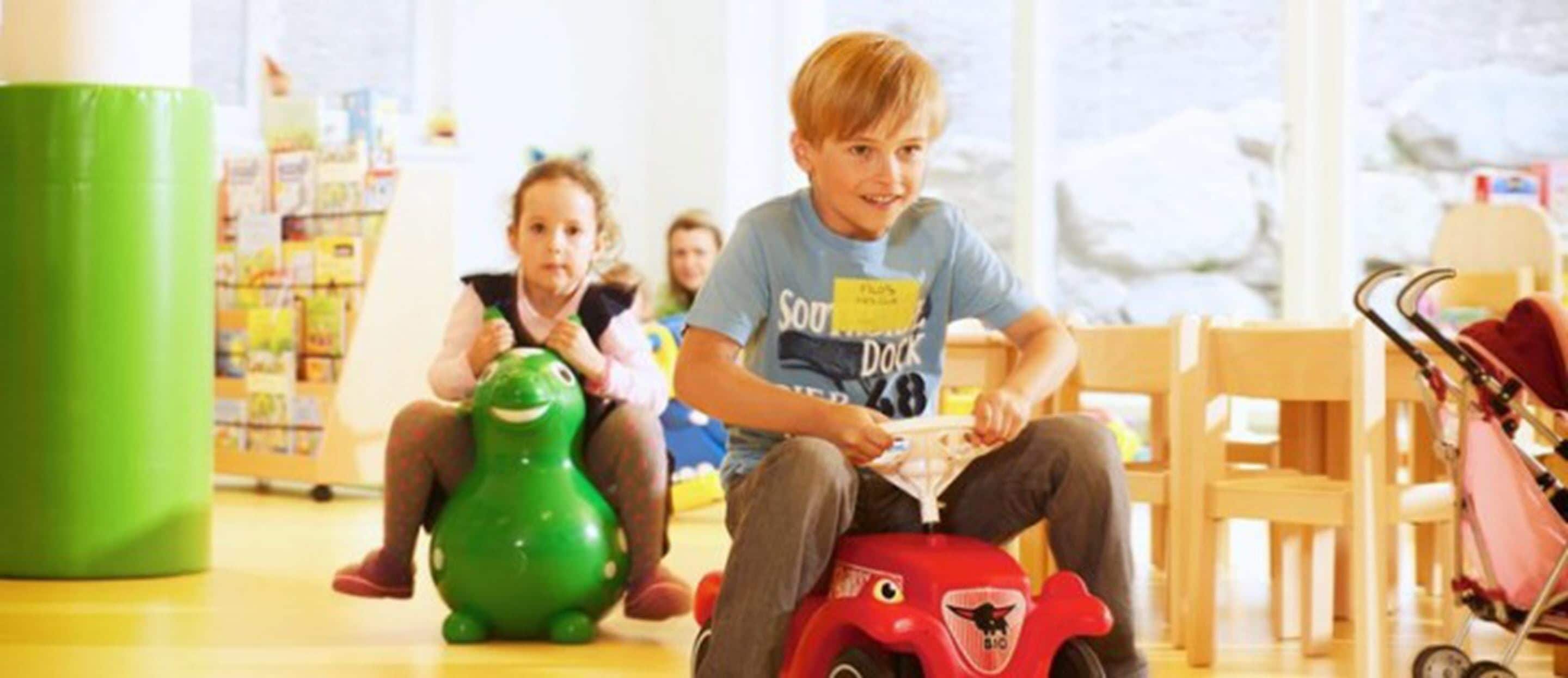 A child on a red toy vehicle in a brightly lit indoor space with wooden furniture and a green pillar.