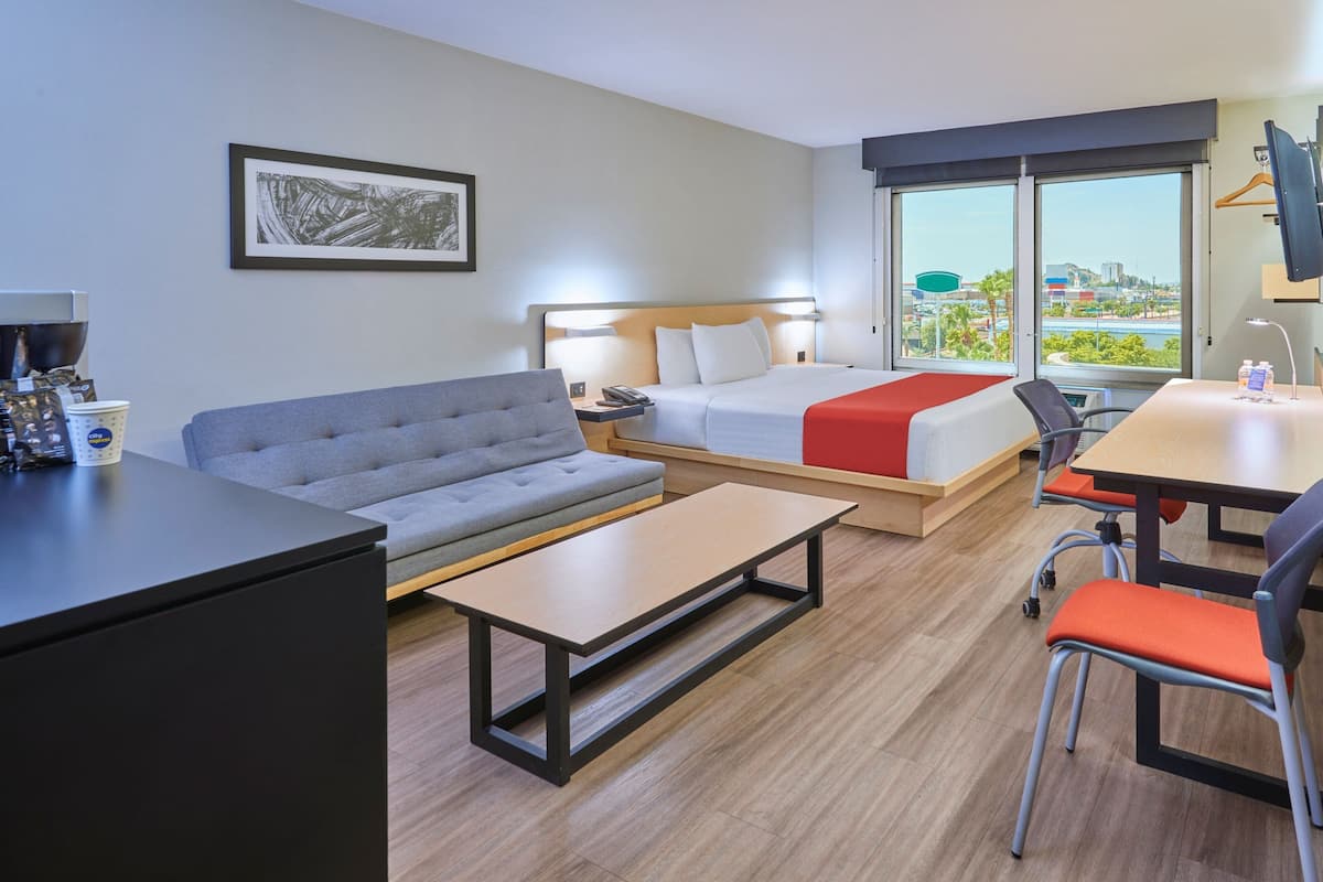 A modern hotel room with a bed, sofa, desk, and a view of the cityscape through large windows.