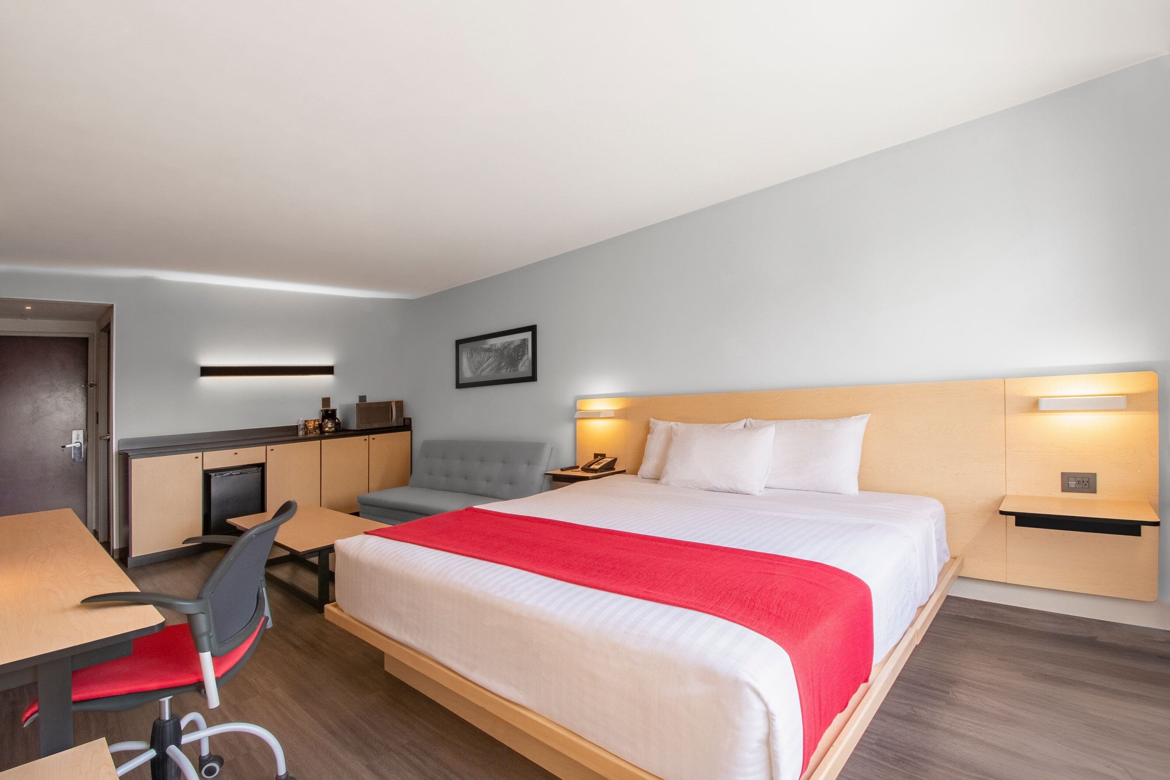Quarto de hotel com cama, escrivaninha, cadeira e uma pequena cozinha compacta.