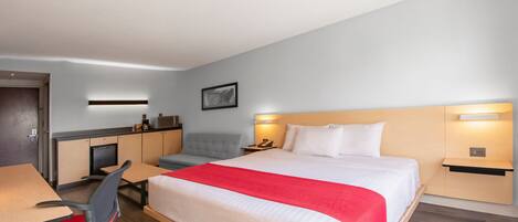 Quarto de hotel com cama, escrivaninha, cadeira e uma pequena cozinha compacta.