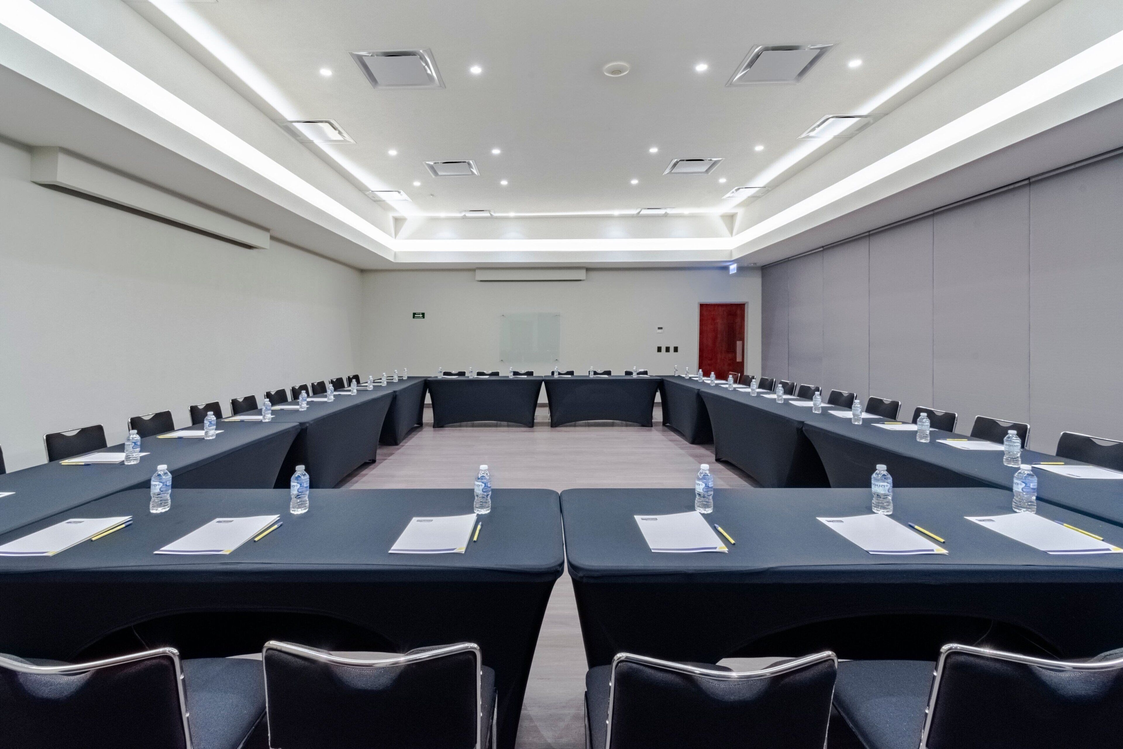 Un salón de conferencias con una larga mesa, sillas, botellas de agua y documentos.
