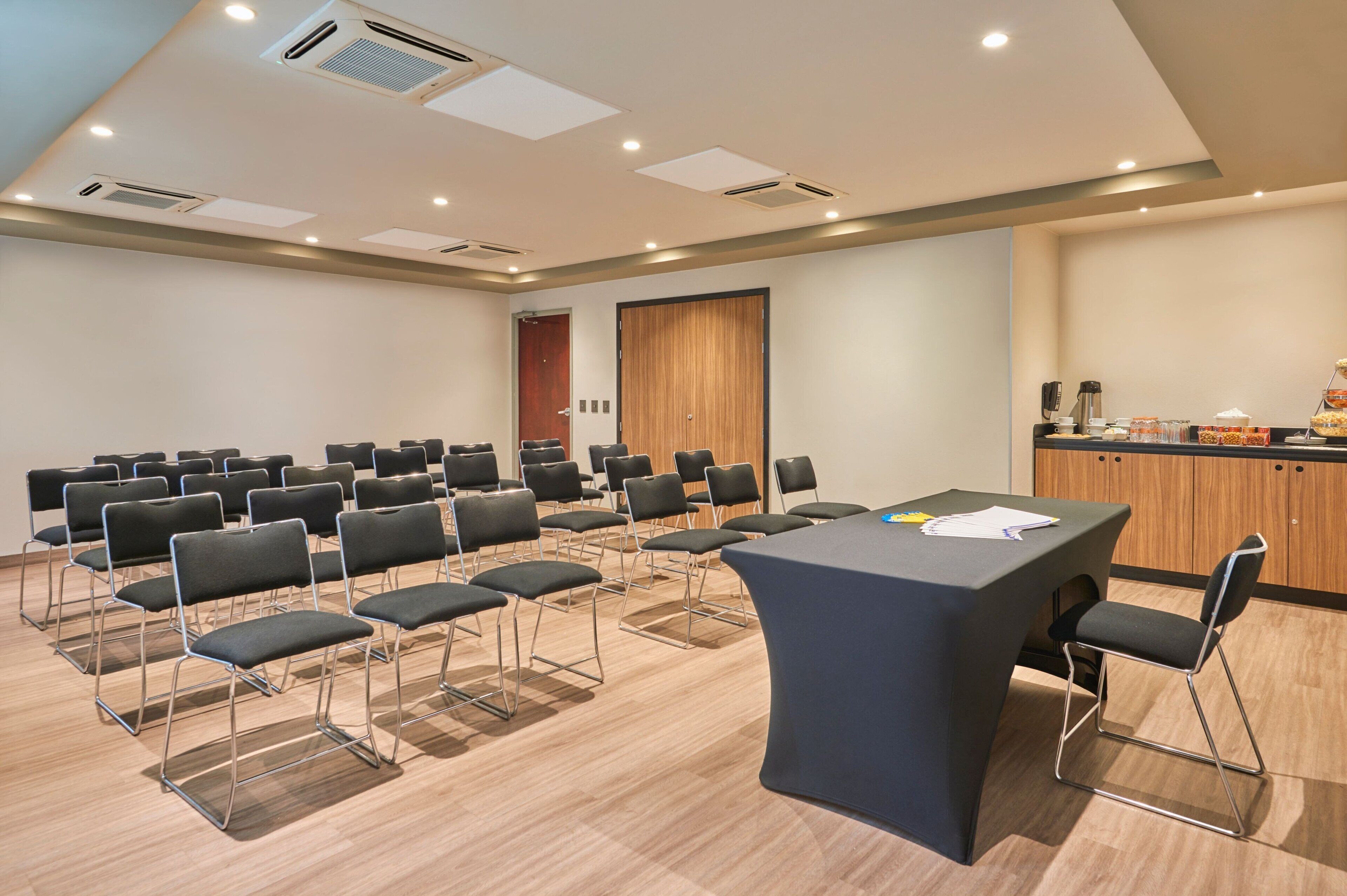 Un salón de conferencias con una mesa larga y varias sillas, una puerta de madera y una zona de kitchenette.