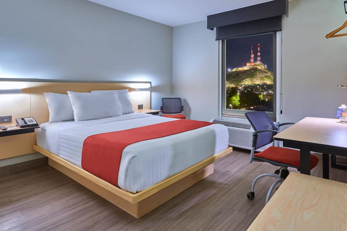 A hotel room with a bed, a desk, a chair, a nightstand, and a window with a view of a tower.