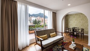 Uma sala de estar moderna com sofá, mesa de jantar e cadeiras, janelas amplas com cortinas e vista para prédios e montanhas.
