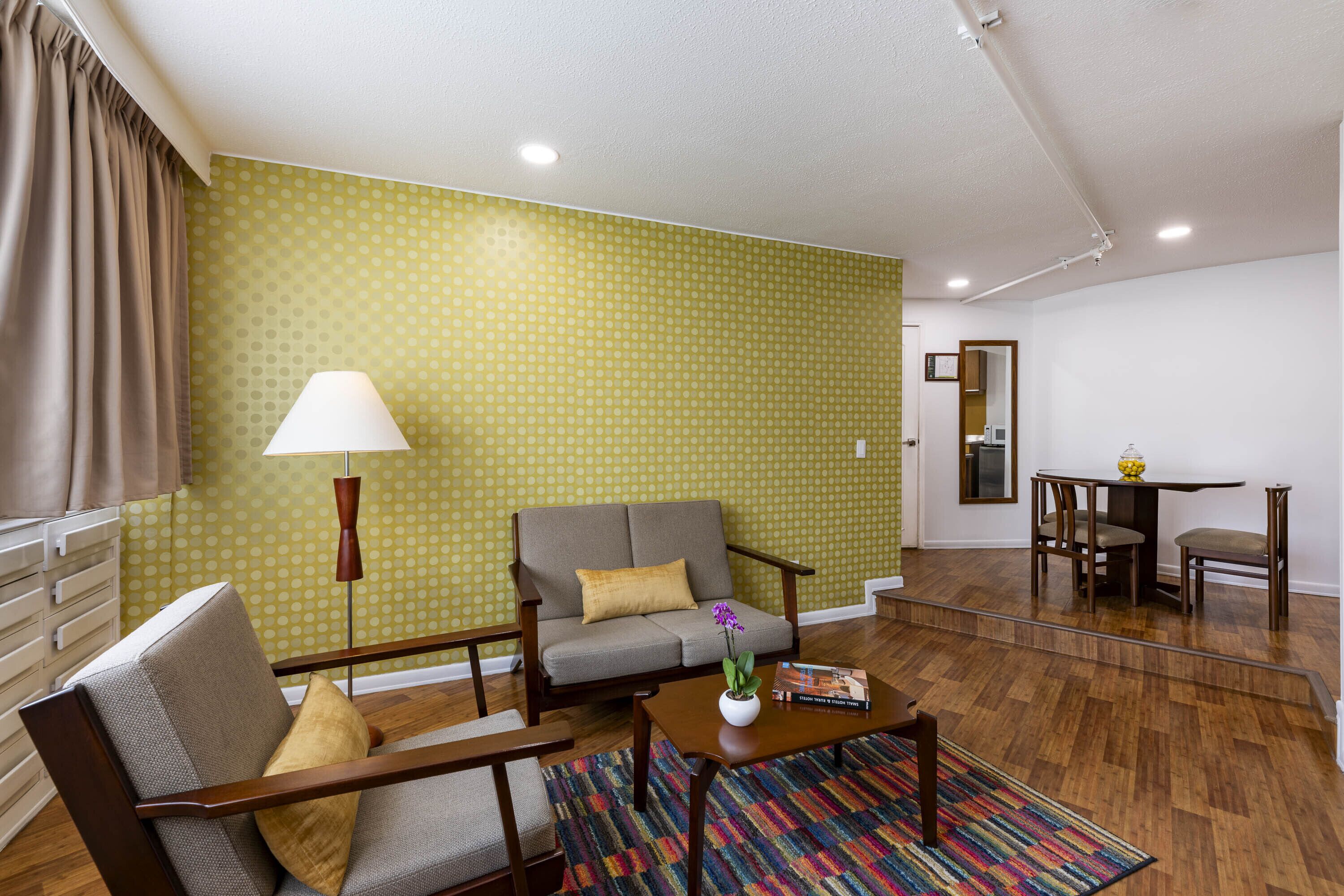 A living room with a sofa, armchair, coffee table, and a lamp. There is a patterned wall and a dining area in the background.