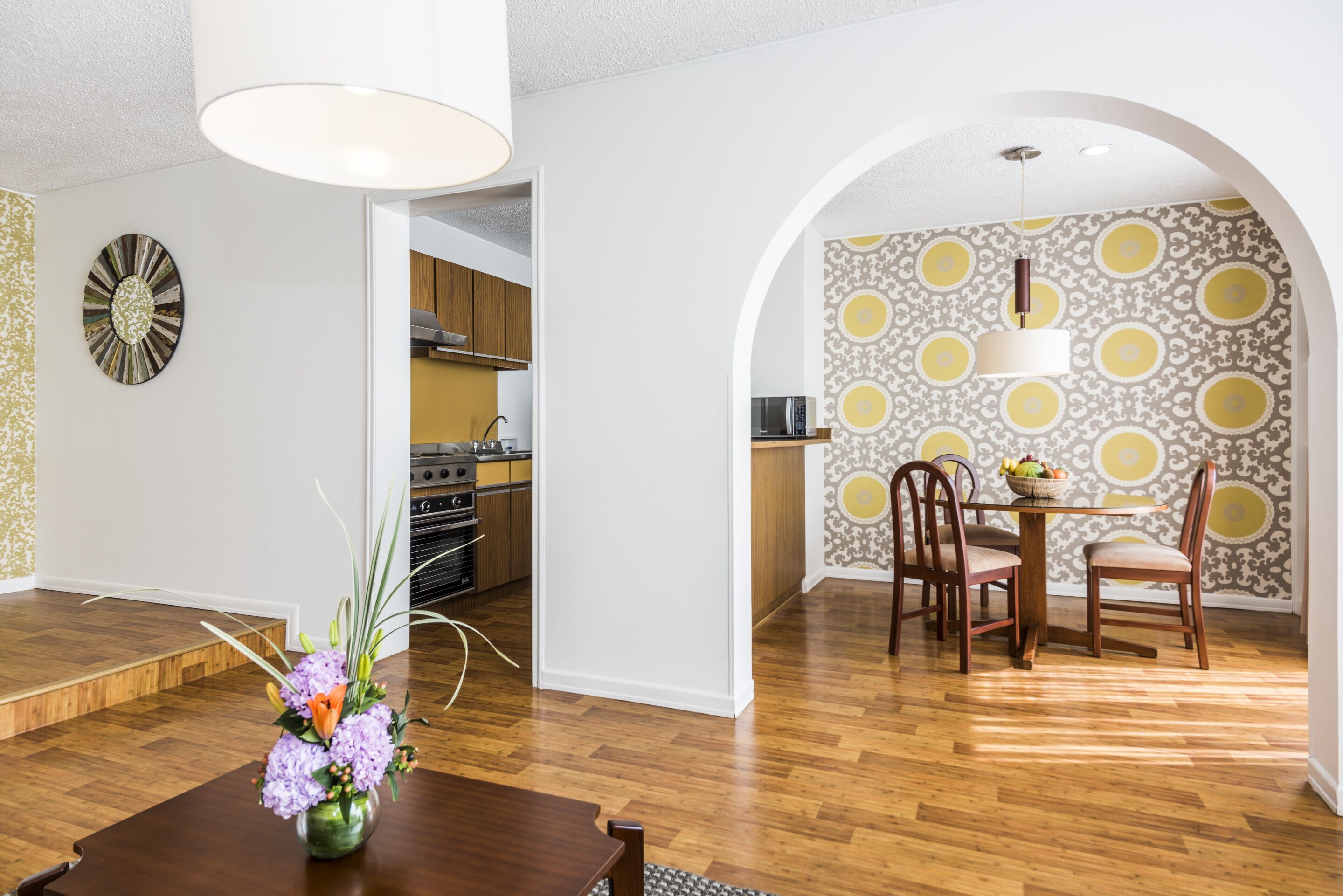 A dining area with a wooden table, chairs, and a vase of flowers. There is a kitchen in the background with wooden cabinets and a microwave.