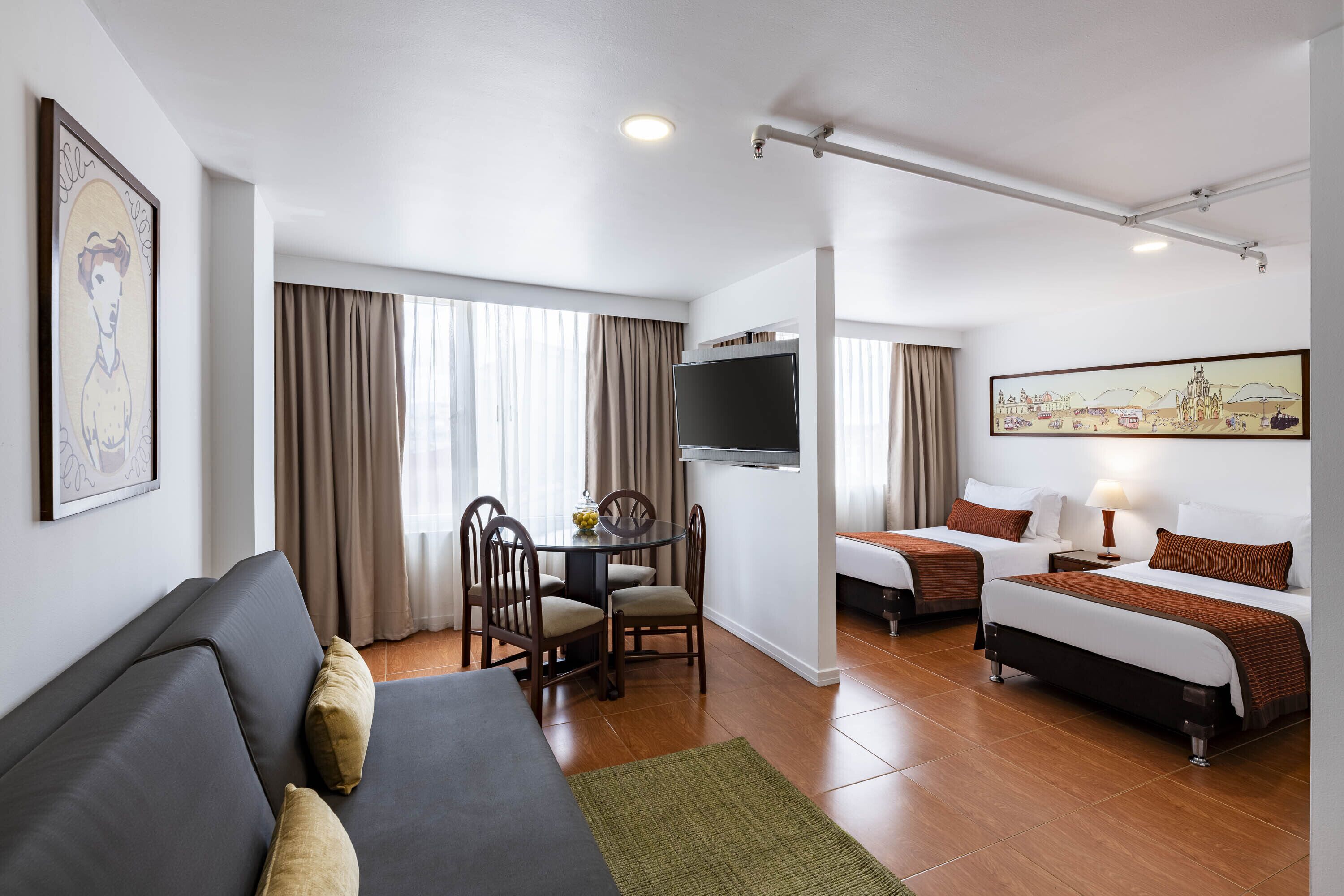 A hotel room with a sofa, two single beds, a dining table with chairs, a flat-screen TV, and artwork on the walls.