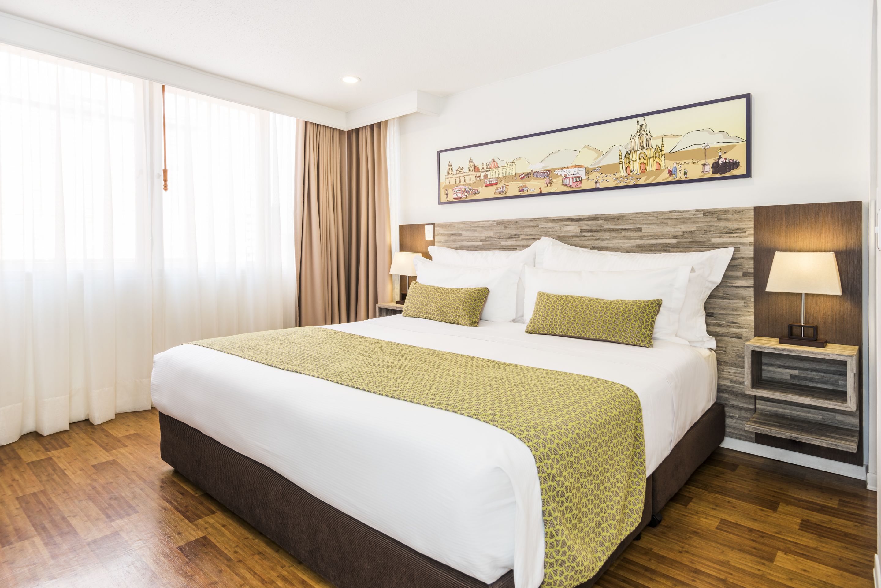 Quarto de hotel com uma cama grande, cabeceira de madeira, mesa de cabeceira com abajur e um quadro sobre a cama.