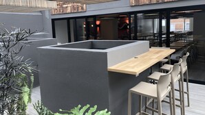 Uma área de estar moderna ao ar livre, com estrutura de concreto, teto de ripas de madeira e banquetas de bar.