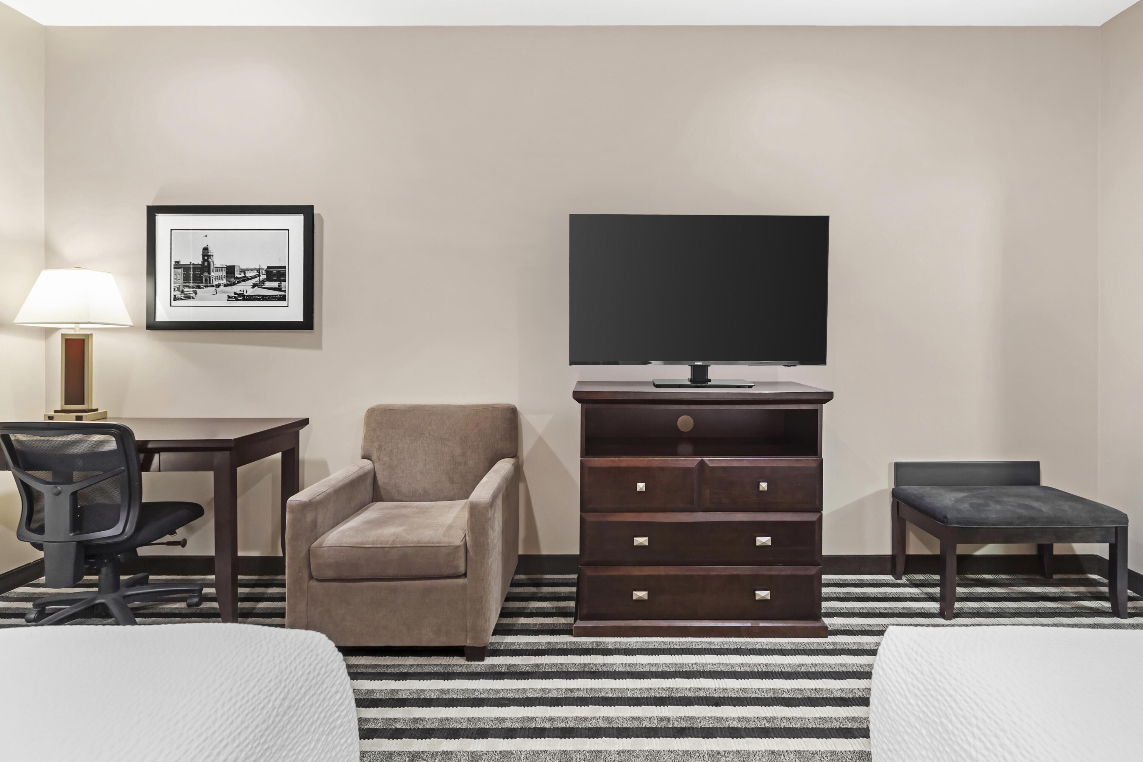 Una habitación de hotel con cama, escritorio, silla, cómoda, televisor y un cuadro en la pared.
