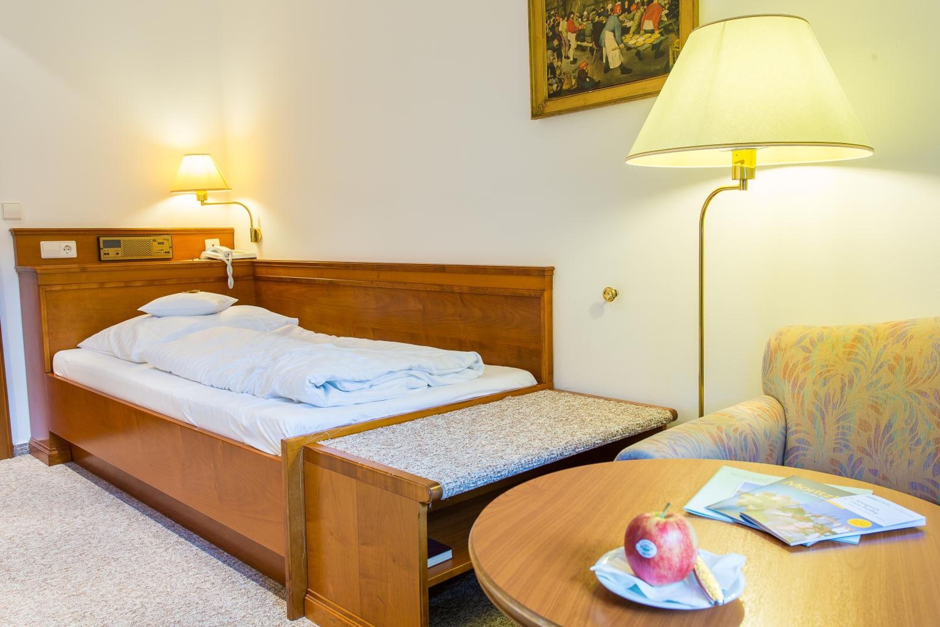 Ett hotellrum med en säng, en sänglampa, ett litet bord med en bok och ett äpple samt en stol.