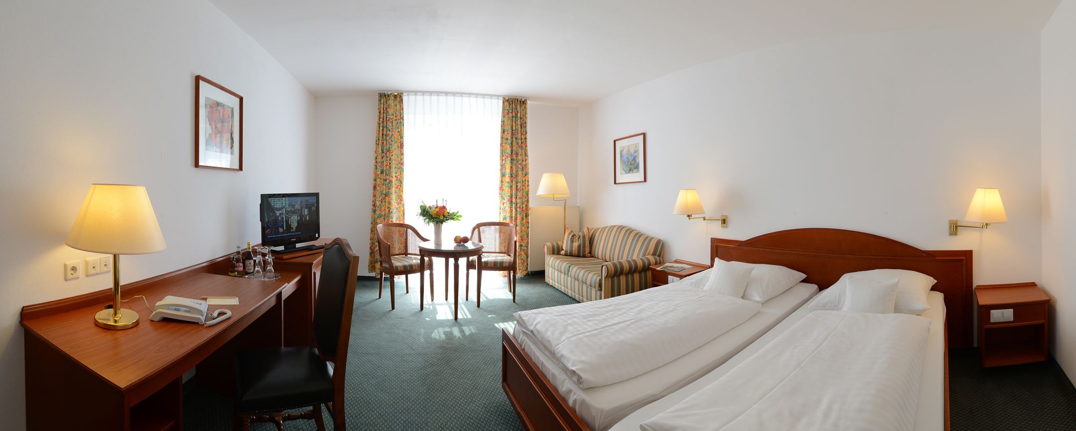 A hotel room with a bed, desk, chair, TV, lamps, and a window with curtains.