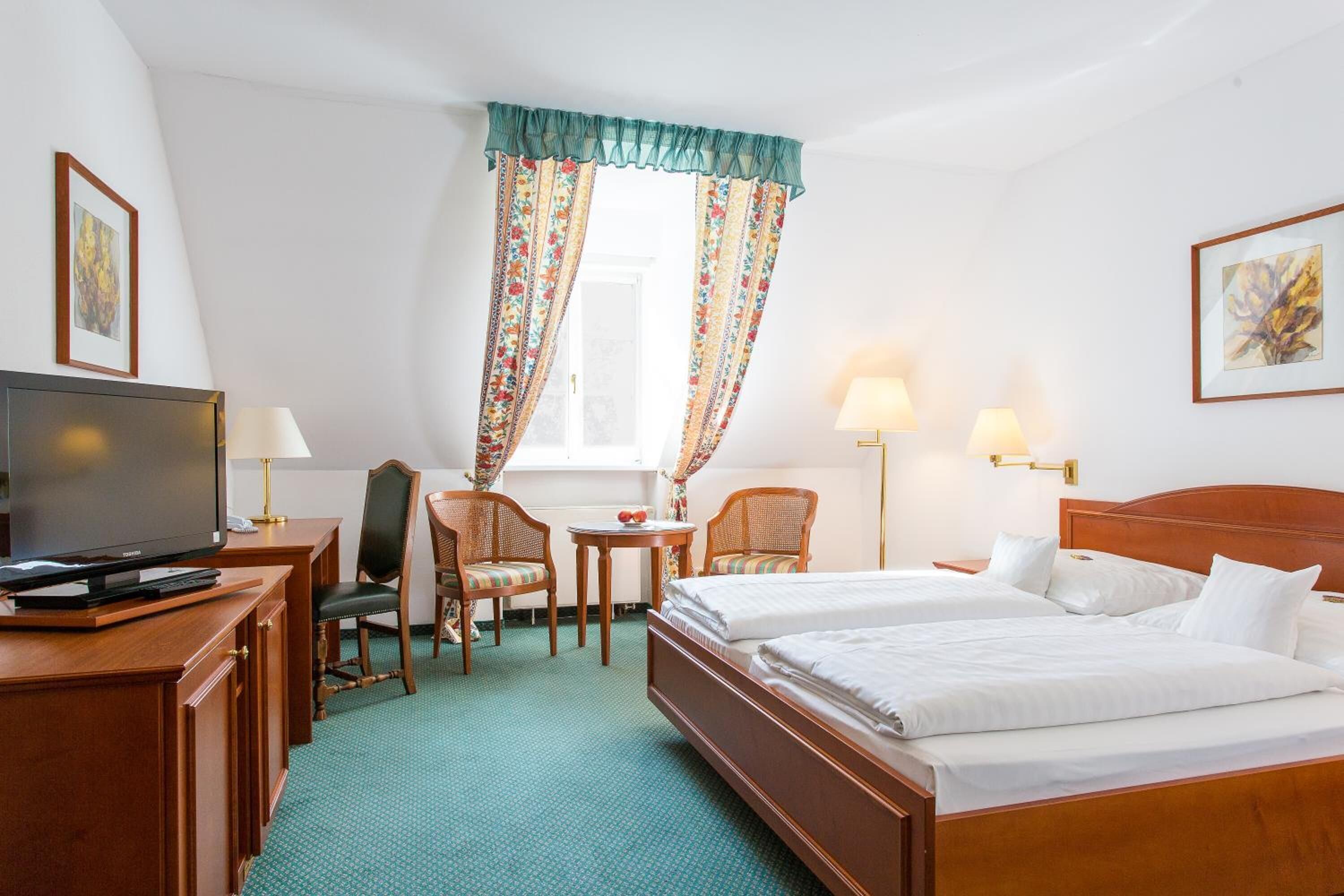 Een hotelkamer met een bed, twee stoelen, een bureau met een televisie en een raam met gordijnen.