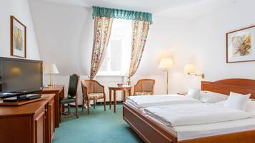 A hotel room with a bed, two chairs, a desk with a television, and a window with curtains.