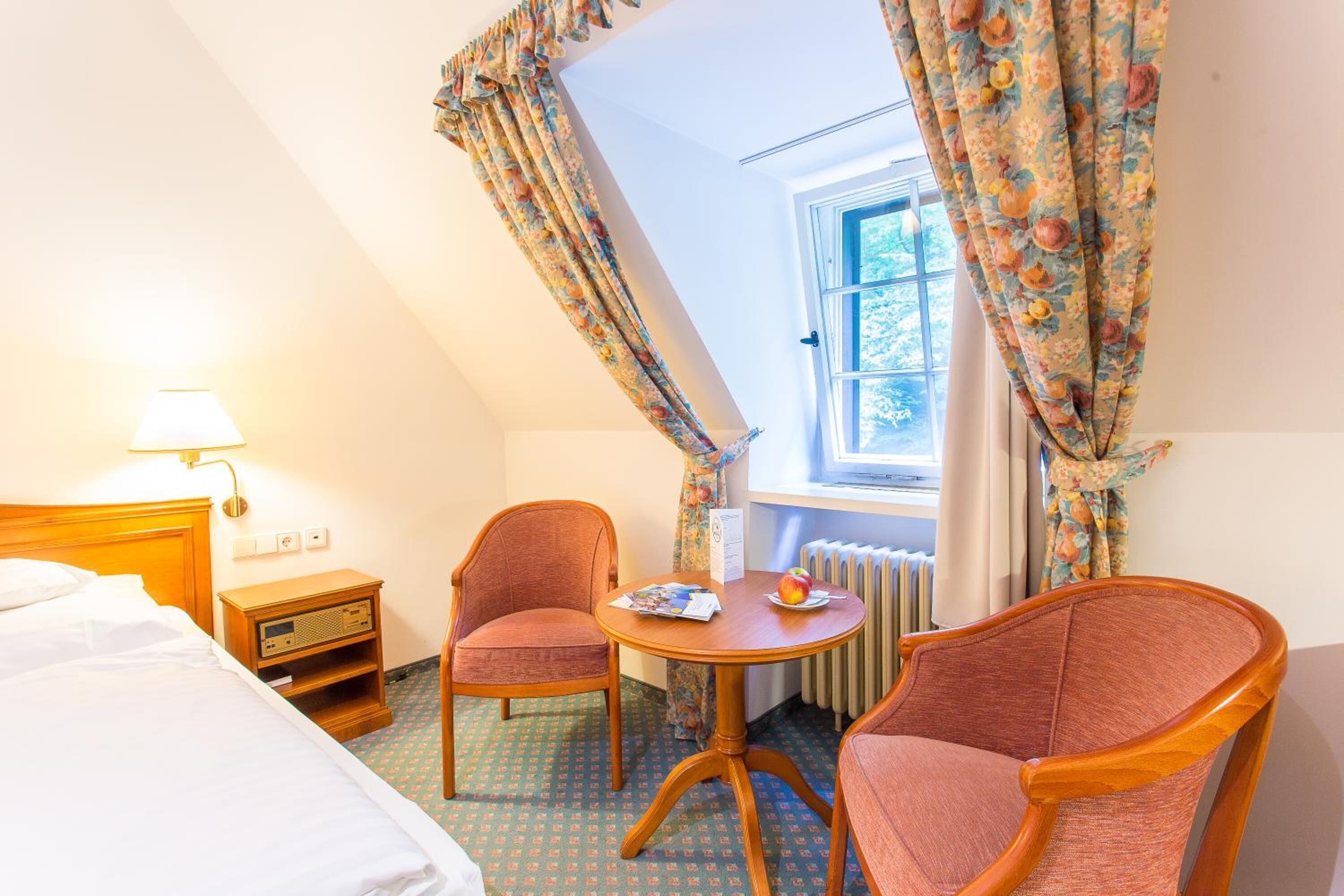 A hotel room with a bed, a nightstand, a table with a chair, and a window with curtains.
