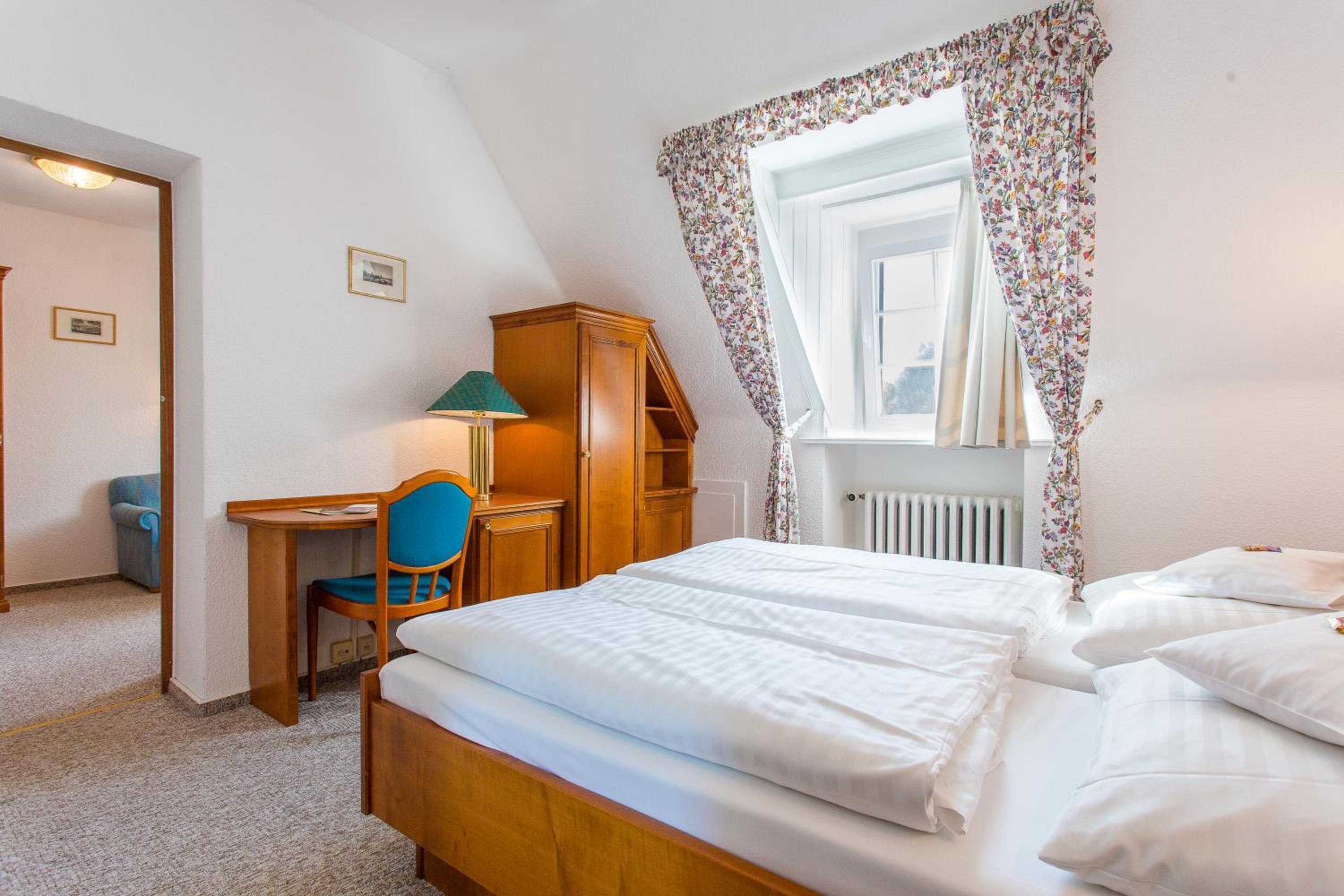 Ein Schlafzimmer mit einem hölzernen Bett, einem Schreibtisch mit Stuhl und einem Fenster mit blumigen Vorhängen.