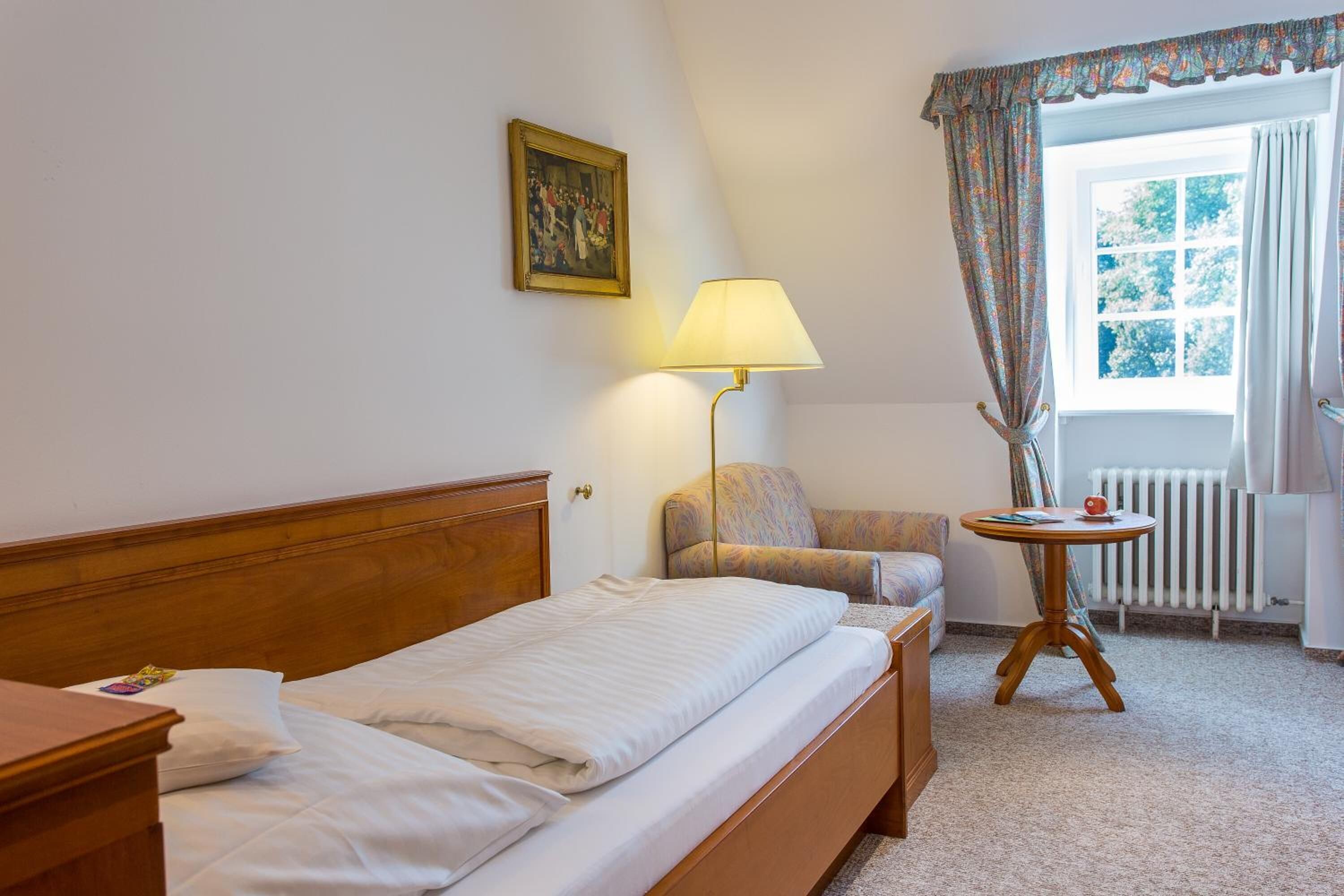 Habitación de hotel con cama, sillón, mesa, lámpara, radiador y ventana con cortinas.