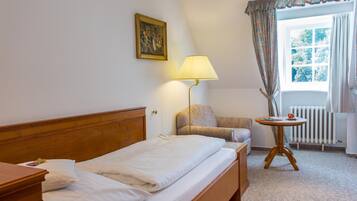 Une chambre d’hôtel avec un lit, une chaise, une table, une lampe, un radiateur et une fenêtre avec des rideaux.