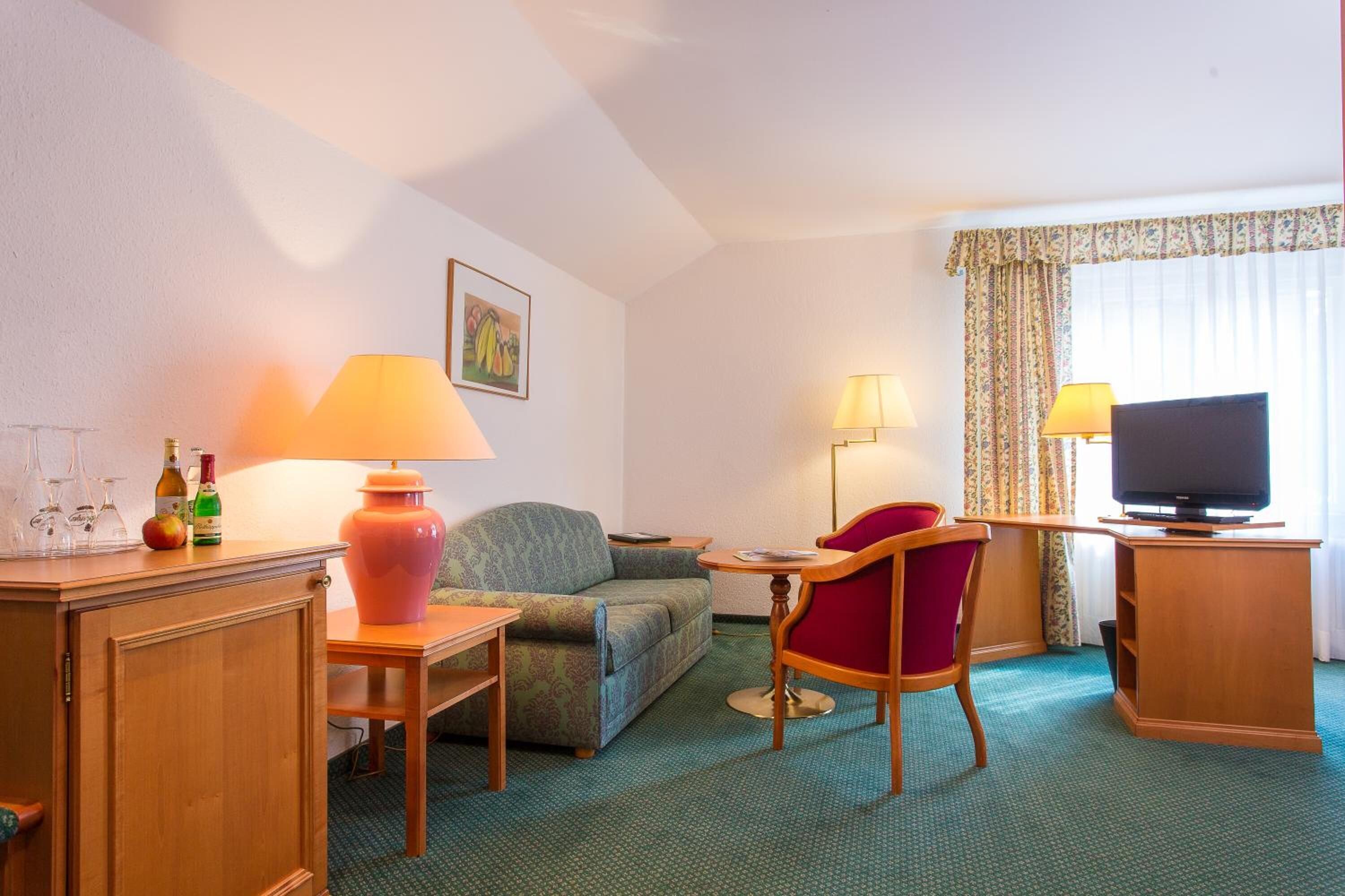 A hotel room with a sofa, a red chair, a television, a table with a lamp, and a cabinet with wine glasses and bottles.