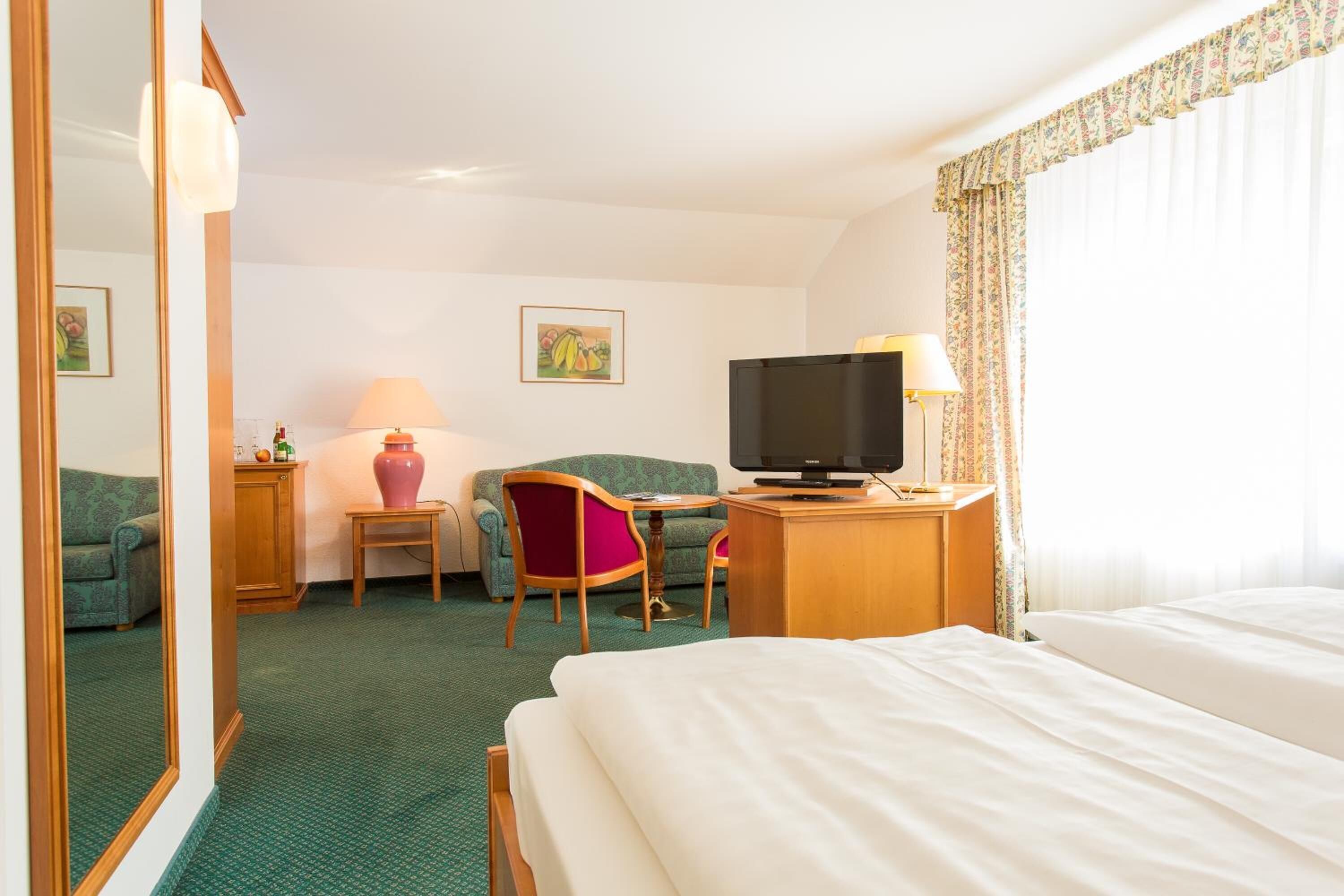 A hotel room with a bed, a desk with a television, a chair, a small table with a lamp, and a mirror.
