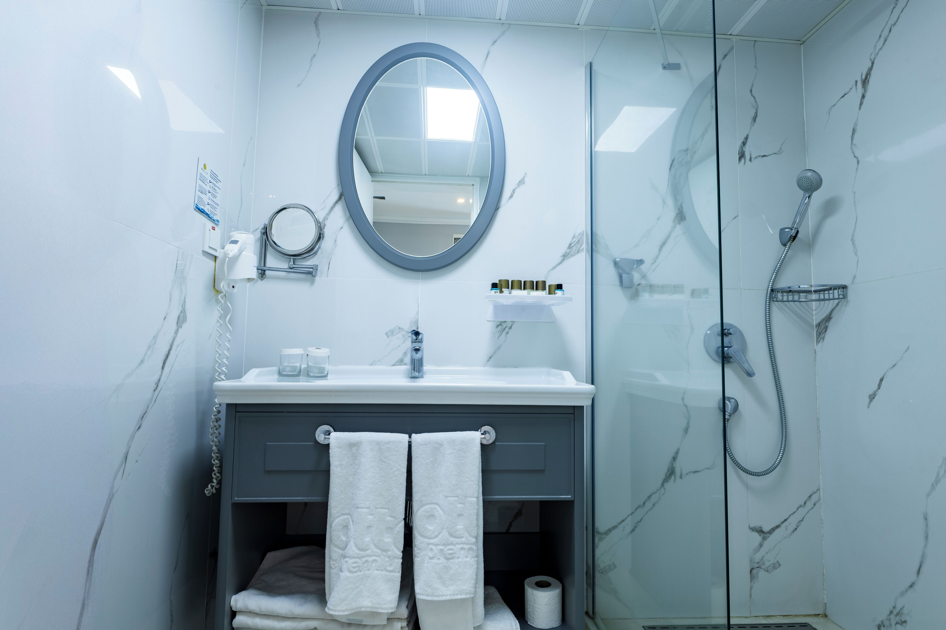 Un baño con pared de mármol, un espejo redondo, un lavamanos con grifo, una ducha con cabezal de ducha de mano y dos toallas colgadas en un perchero.