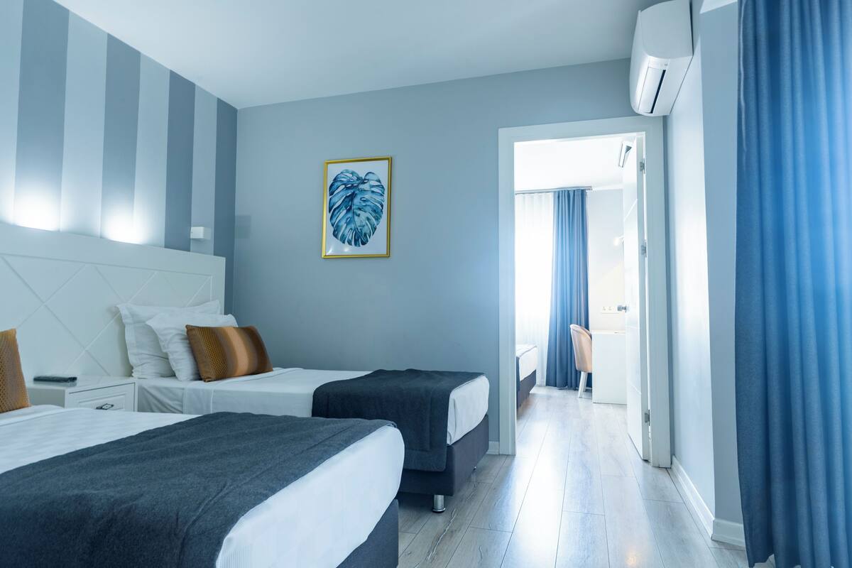 A hotel room with two beds, a large window with curtains, a wall-mounted air conditioner, and a framed picture.