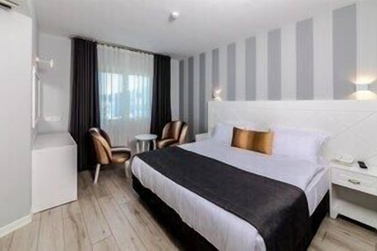 A hotel room with a large bed, a bedside table, a chair, a small table, and a window with curtains.