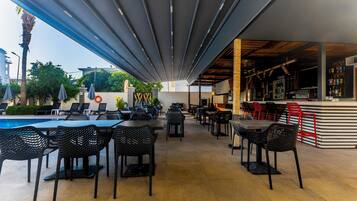 An outdoor cafe with tables and chairs, a pool, and a bar area.