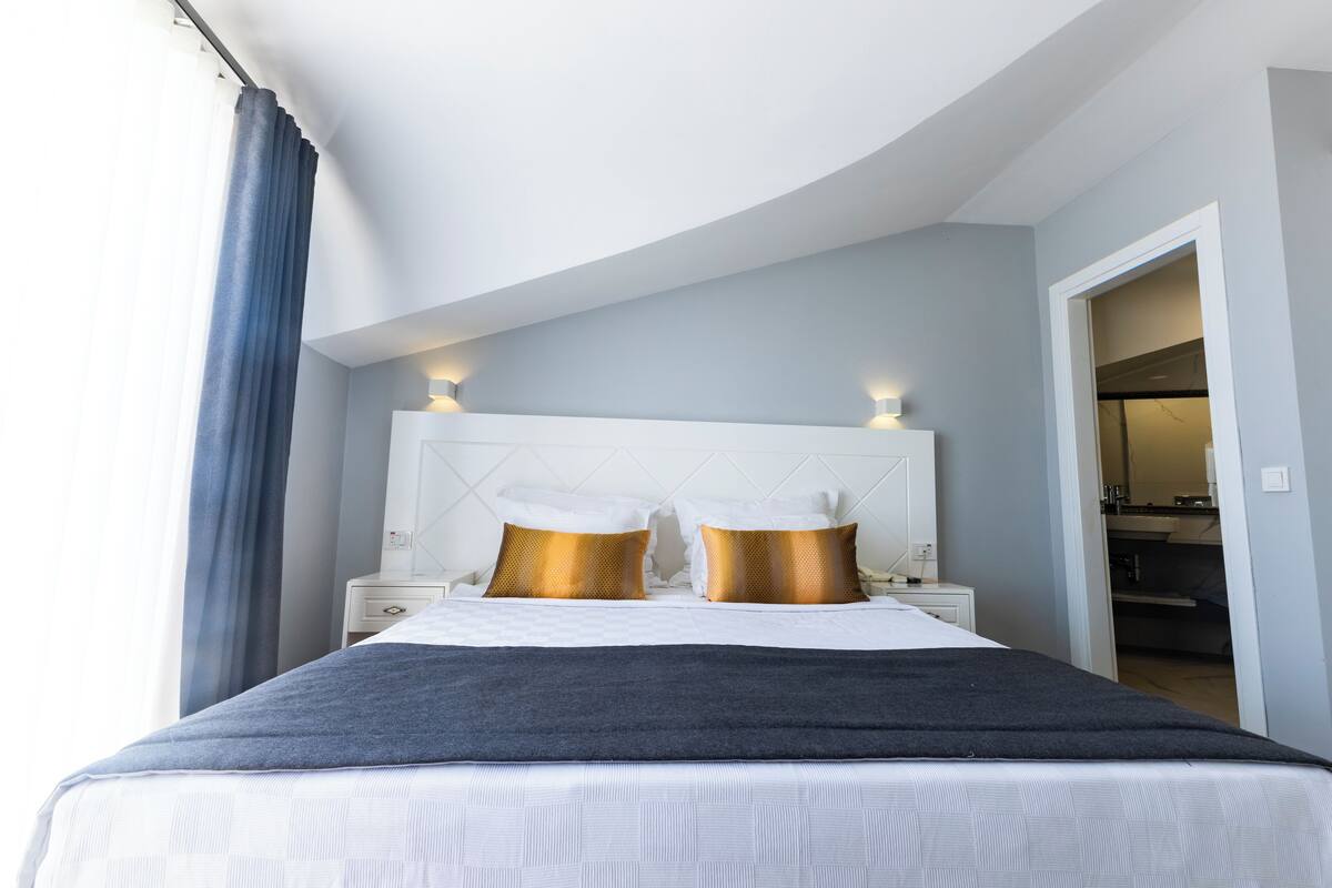 A modern hotel room with a curved ceiling, a large bed with white bedding and gold accent pillows, a dark grey throw blanket, and a white nightstand with a lamp.