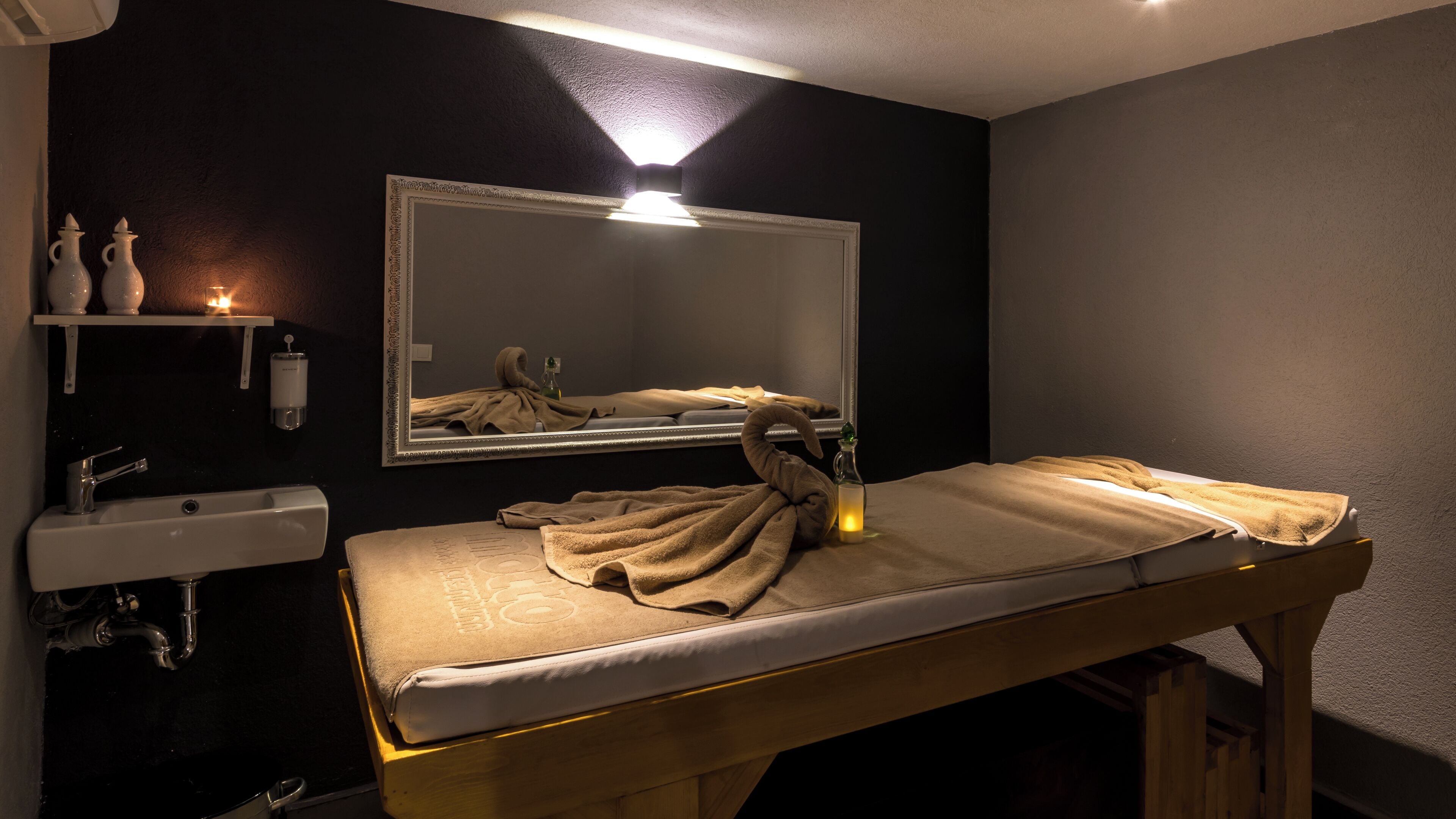 A spa room with a wooden massage table, a sink, a mirror, and a shelf with bottles.