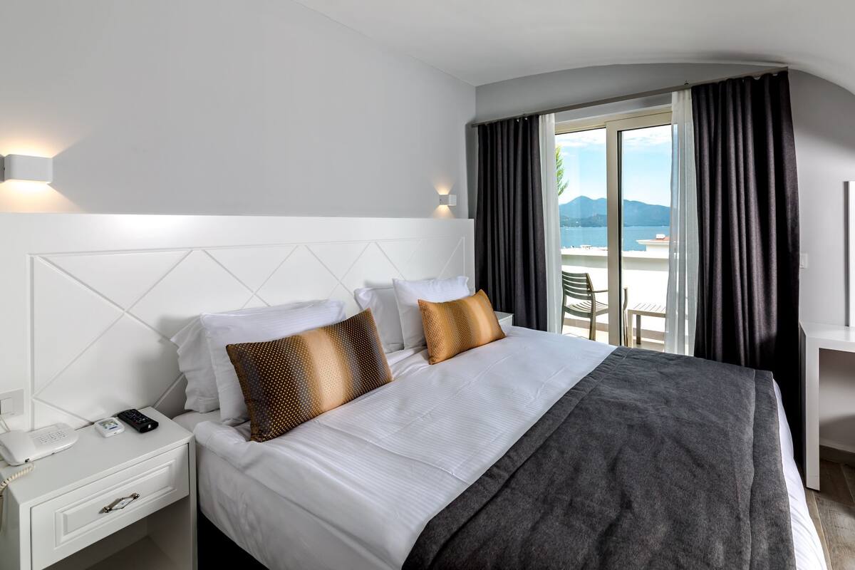 A hotel room with a bed, a bedside telephone, a desk with a chair, and a view of the outdoors.
