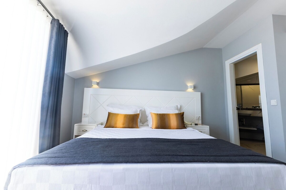 A hotel room with a curved ceiling, a bed with white bedding and two gold pillows, a nightstand with a lamp, and a view of a bathroom and closet.
