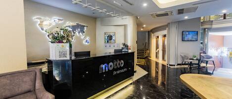 A hotel reception area with a black reception desk labeled "motto Premium," a world map light installation, a seating area with a wooden table, and a modern interior design.