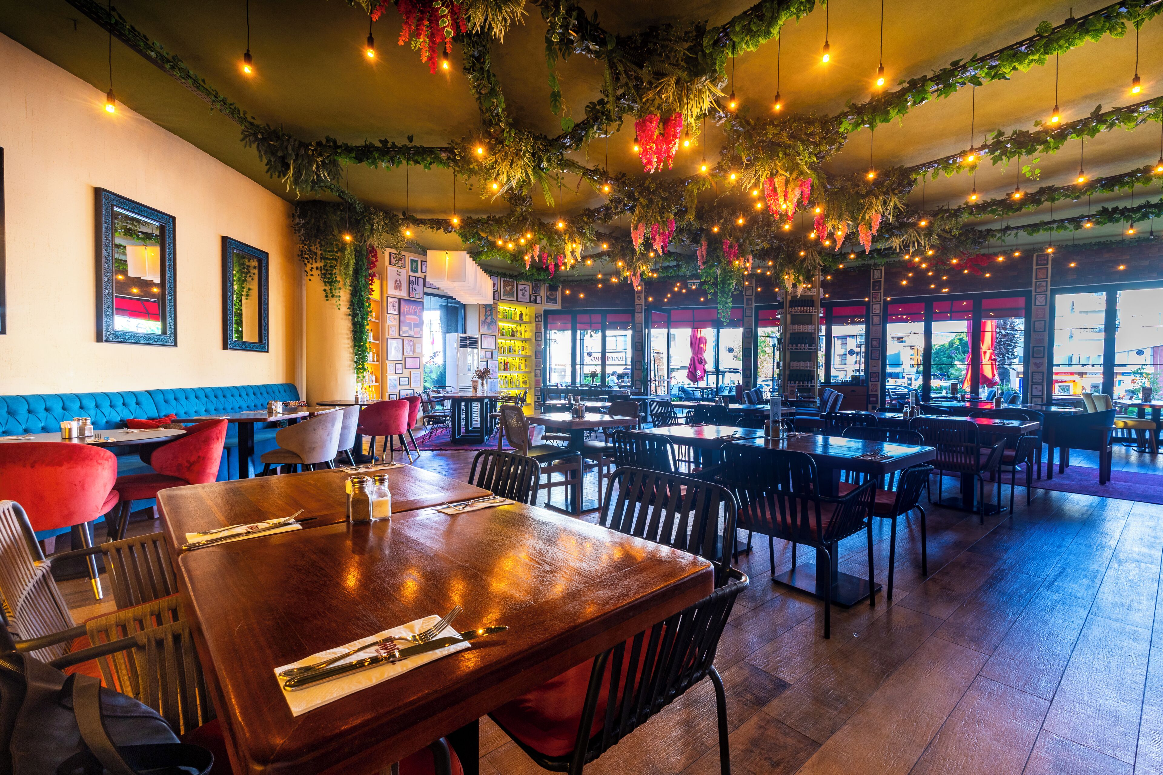Een restaurant met houten tafels en stoelen, blauwe gestoffeerde zitjes en hangplanten met verlichting.