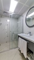 Un baño moderno con cabina de ducha de vidrio, pared con azulejos de mármol, espejo redondo y lavabo blanco con una toalla colgada en la puerta.