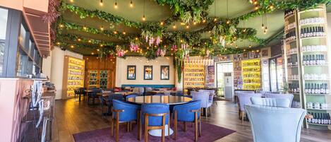 Un restaurant avec une table ronde entourée de chaises bleues, un coin bar avec des étagères remplies de bouteilles, et un plafond décoré de plantes et de luminaires suspendus.