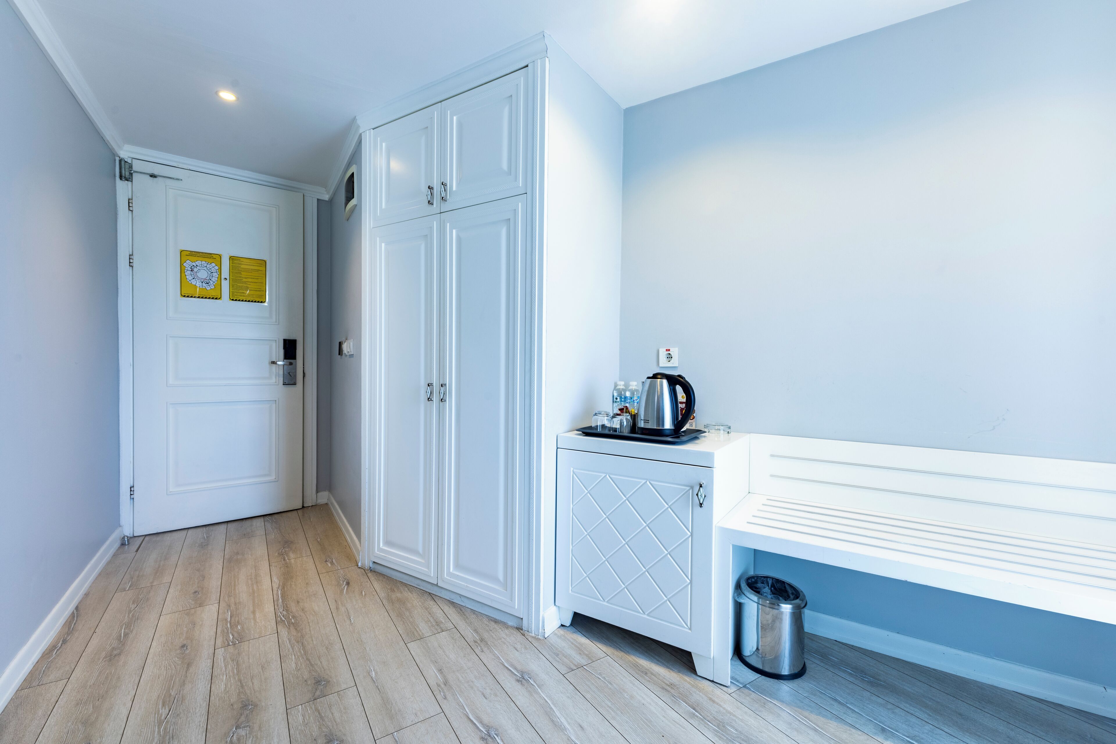 Un pasillo con una puerta blanca, un mueble blanco con una bandeja y un bote de basura.